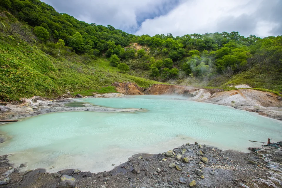 Steam rising from pale blue geothermal water surrounded by forest in Noboribetsu