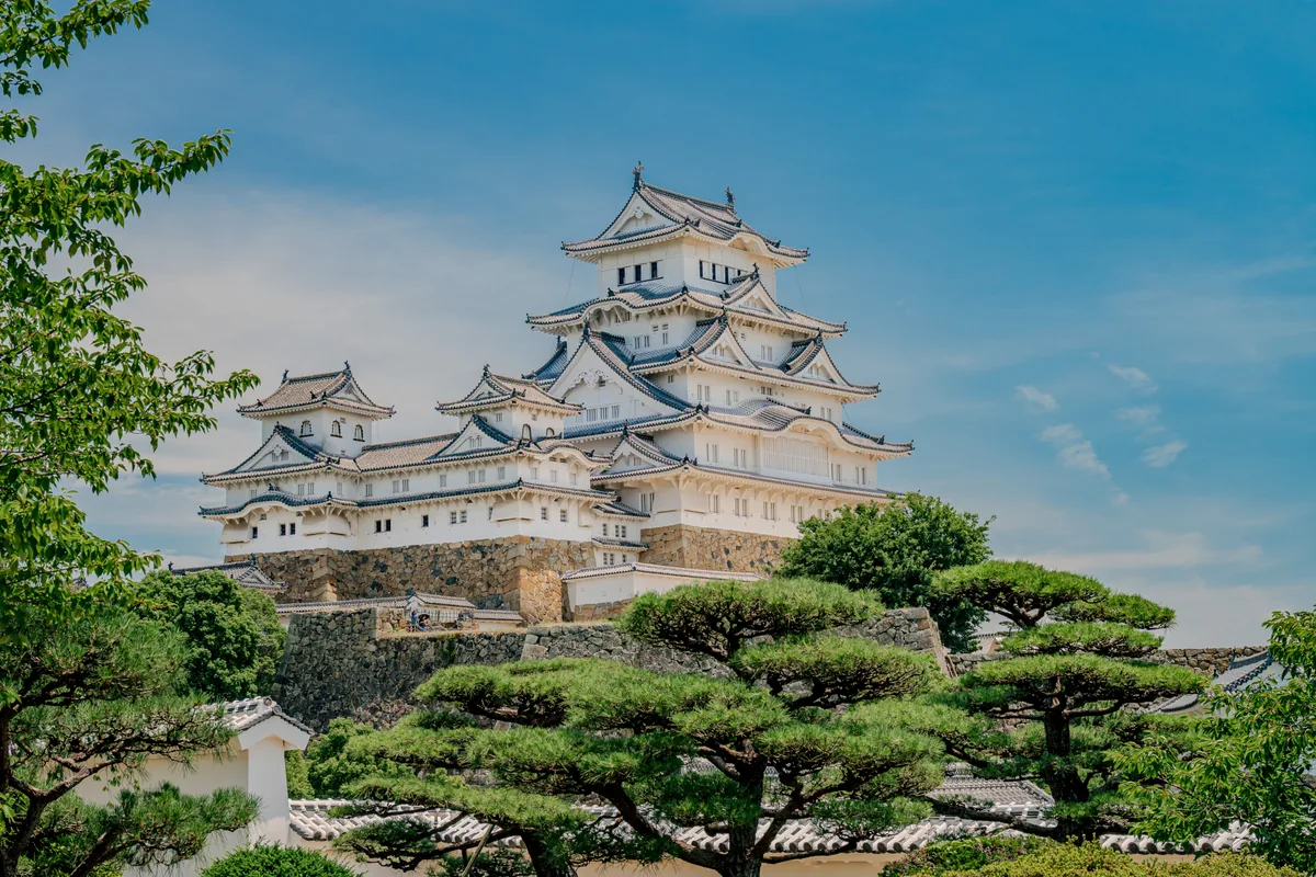 Himeji Castle’s white towers and stone walls rising above green trees under a blue sky