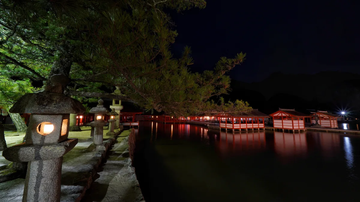 Itsukushima Shrine illuminated across the water at night, with glowing stone lanterns in the foreground