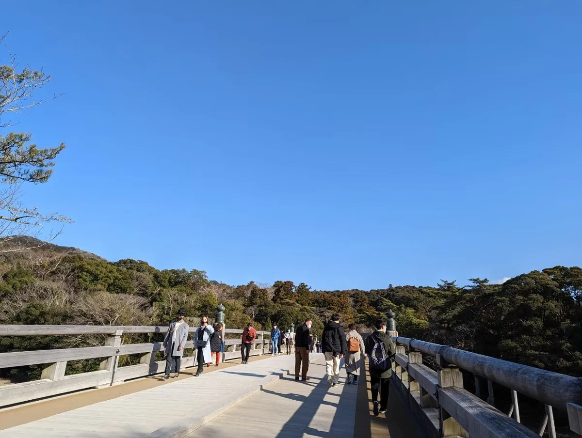 Visitors crossing the wooden Uji Bridge toward wooded hills beneath a blue sky