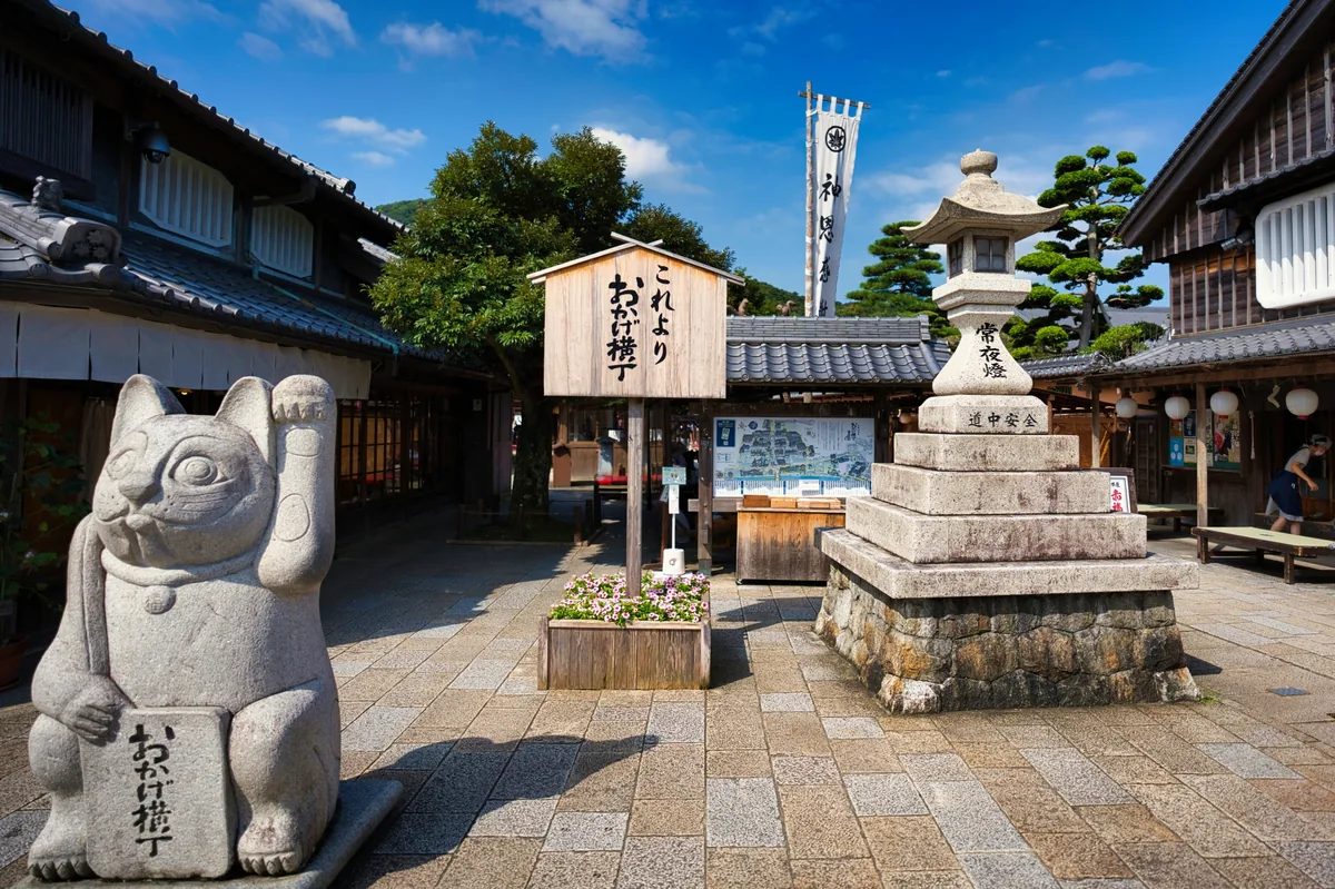 A stone beckoning cat and lantern beside an Okage Yokocho entrance sign