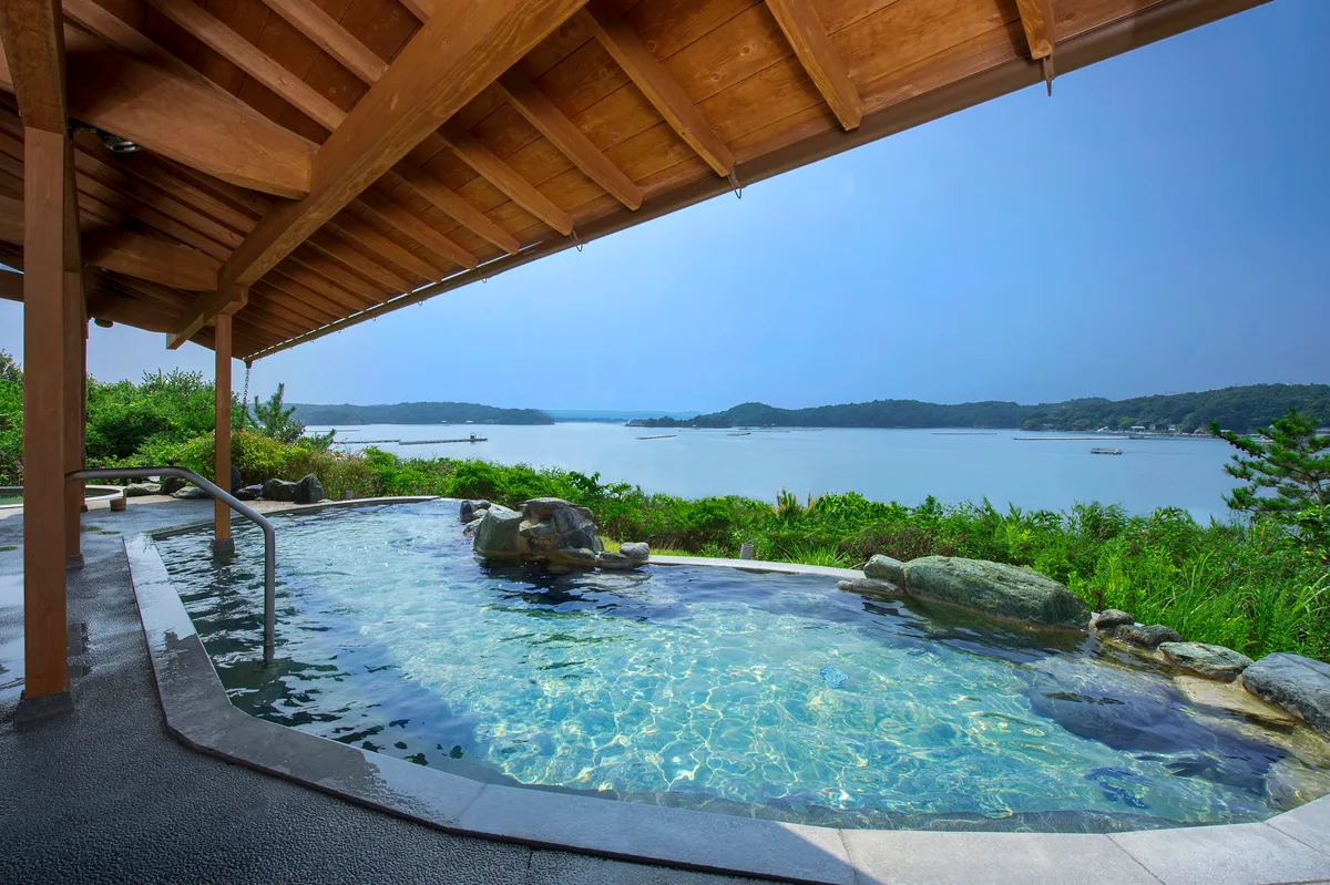 Asanagi-no-Yu garden bath beneath a wooden roof, facing blue water and the wooded shores of Ago Bay