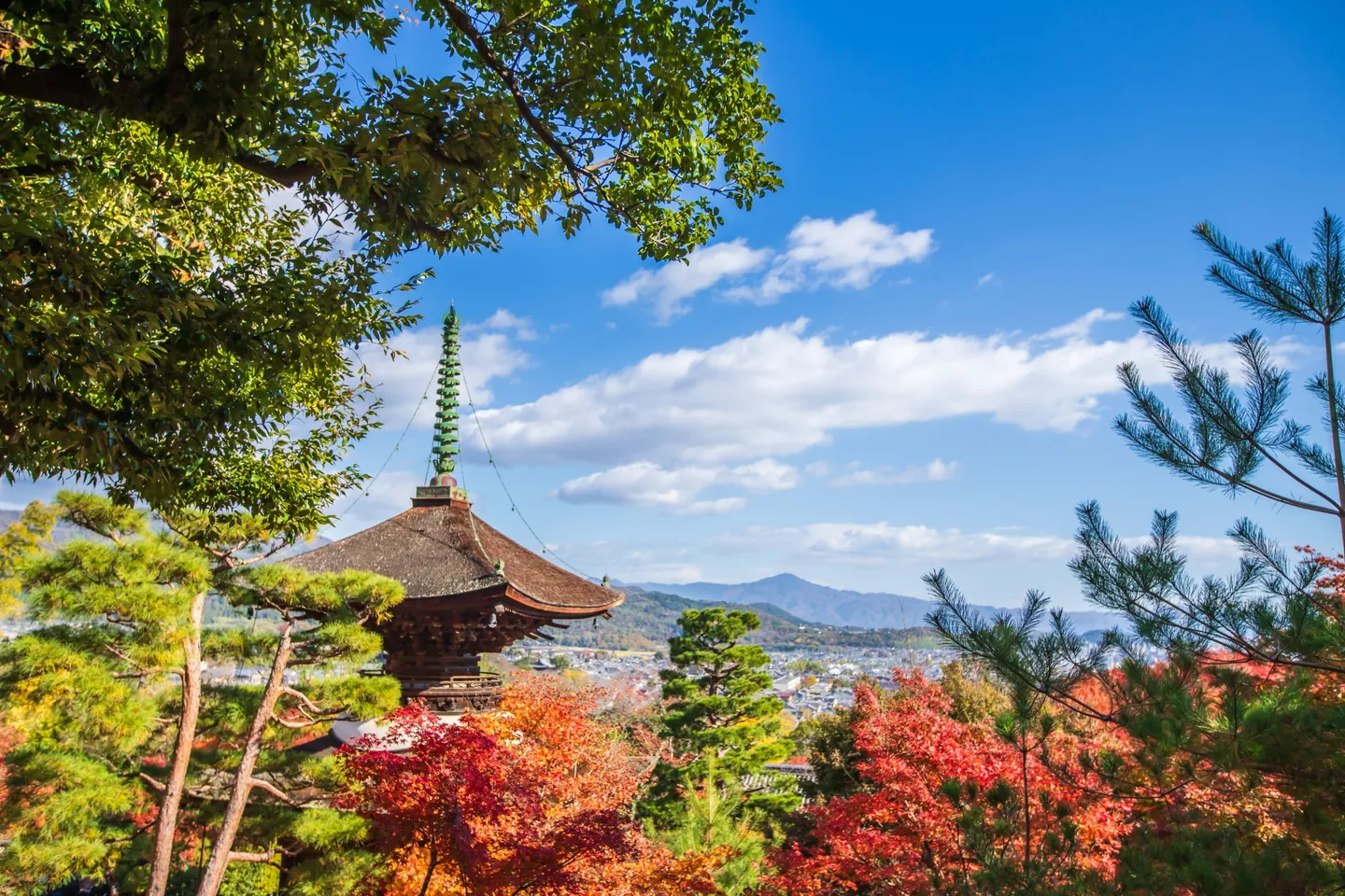 The Tahoto pagoda rises above autumn maples at Jojakko-ji, with Kyoto buildings and distant hills beneath a blue sky.