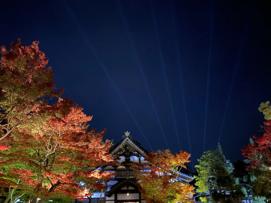 Illuminated autumn leaves frame a temple building beneath a deep blue sky with beams of light at Kodai-ji.