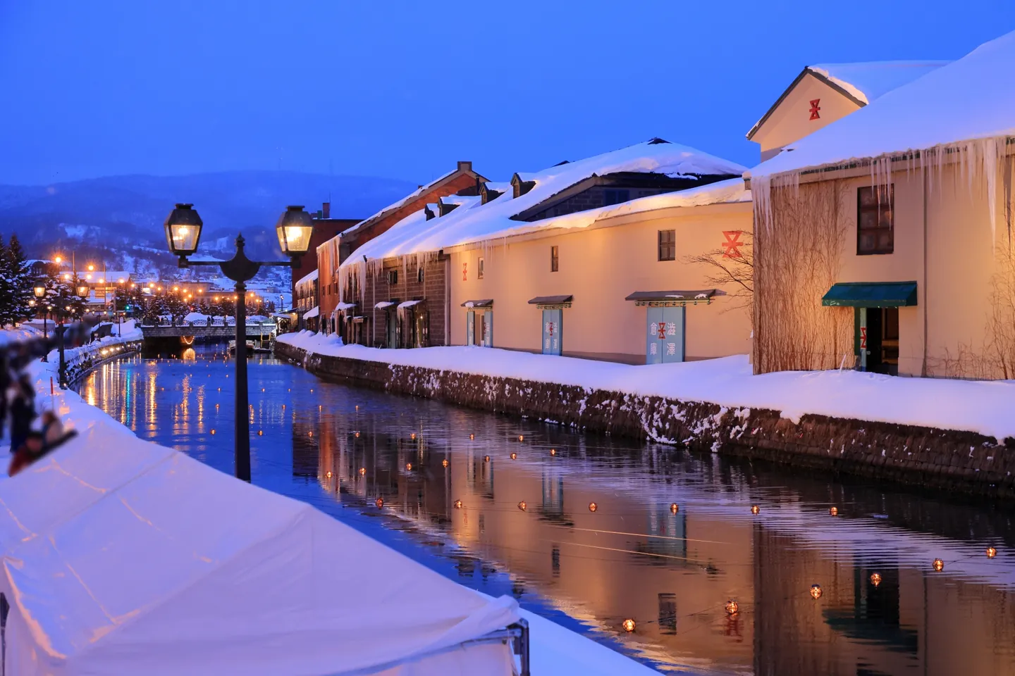 Warm lamps reflected in Otaru Canal at dusk beside snow-covered banks and stone warehouses